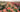 A row of peachy orange roses in a field with a row of yellow roses on the left and red on the right. 