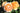 Orange and cream rose blooms on a garden bush with green leaves and soil in the background.