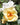 A close-up of a pale yellow rose in full bloom with a bright yellow-orange center and white petals showing from the rose behind it, surrounded by green leaves and a softly blurred background.