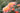 Orange-pink roses with green leaves in a garden setting, featuring a large fully bloomed flower, a bud, and another partially opened bloom against a soft blurred background.